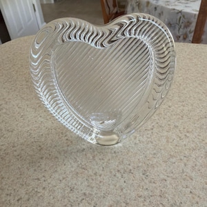May include: Clear glass heart-shaped dish with a textured, wavy border and diagonal lines inside. The dish sits on a light-colored surface. This decorative piece could be used for serving or display.