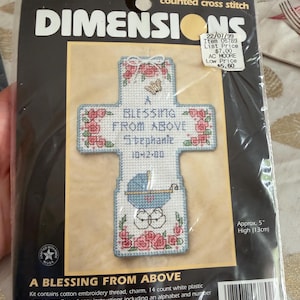 May include: A counted cross stitch kit by Dimensions, featuring a cross design with the text "A Blessing From Above". The cross is decorated with pink roses, a butterfly, and a baby carriage. The kit is approximately 5 inches (13cm) high.