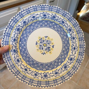 May include: A round ceramic dinner plate with a scalloped edge featuring a white center with a blue and yellow floral medallion. The border displays a blue and white plaid pattern, a band of small blue flowers, and a thin yellow accent line.