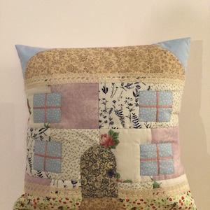May include: A patchwork cushion in the shape of a house. It features a light blue roof, floral and patterned fabrics, and lace trim. The windows are light blue with red trim, and the door has a floral pattern.