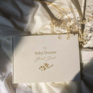 May include: A white guest book with gold lettering that reads "The Baby Shower Guest Book". A small gold leaf design is below the text. The book is lying on a white fabric surface.