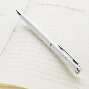 May include: A white pen with a silver clip and a crystal on the end. The pen has the word "love" inscribed on the side. The pen is lying on a lined notebook with the text "What I miss most about you is..." written at the top.