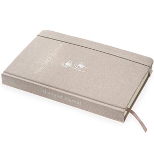 May include: A beige linen-covered journal with an elastic closure and a ribbon bookmark. The front cover features the text "The Grief Journal" and an image of two dandelions.
