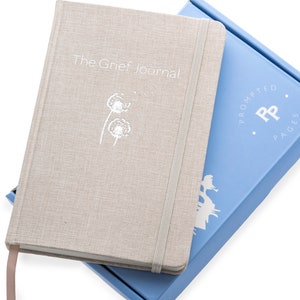 May include: A beige linen journal with a white dandelion design and the text "The Grief Journal". The journal is closed and has a white elastic band. The journal is sitting on top of a light blue box with the text "Prompted Pages" and a white "PP" logo.