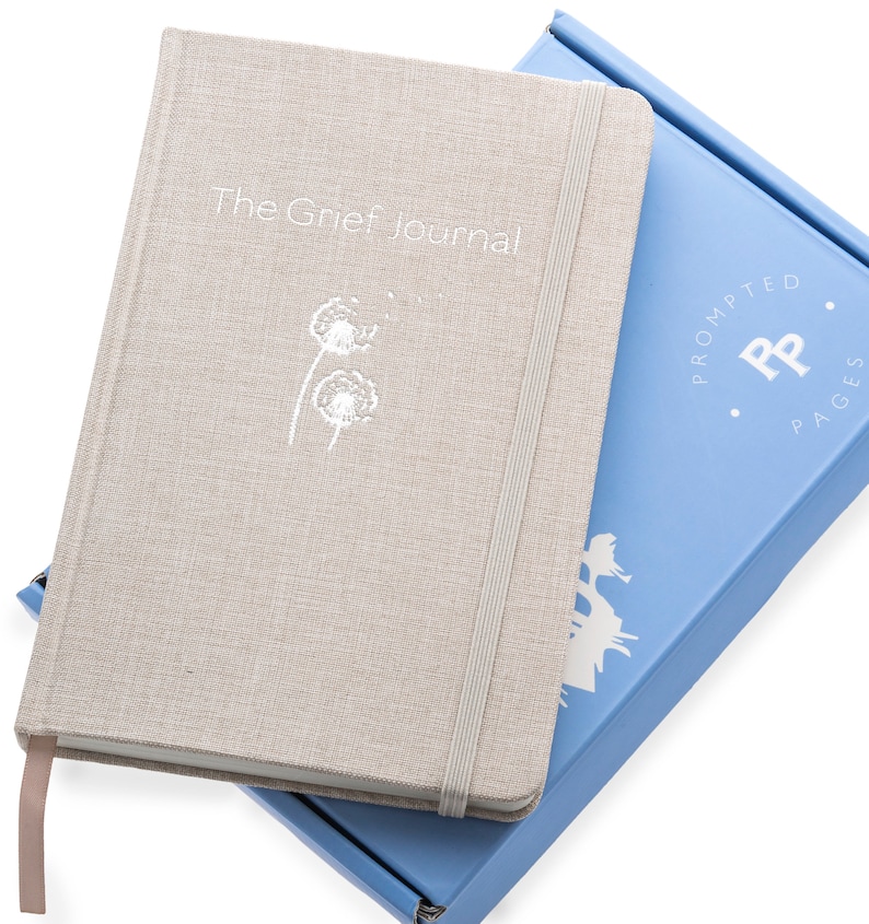 May include: A beige linen journal with a white dandelion design and the text "The Grief Journal". The journal is closed and has a white elastic band. The journal is sitting on top of a light blue box with the text "Prompted Pages" and a white "PP" logo.
