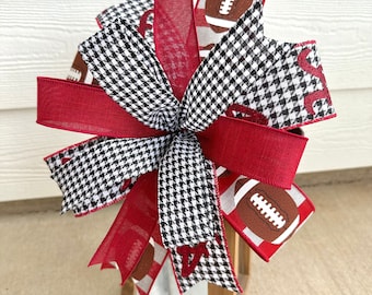 May include: A decorative bow featuring a mix of red, black and white patterned ribbons. The bow is adorned with football-themed ribbon and houndstooth patterns. The bow is placed on top of a wooden lantern.