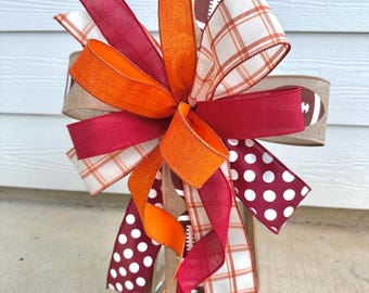 May include: A decorative bow featuring a football theme. The bow has layers of ribbons in orange, burgundy, and white with plaid and polka dot patterns. A football is printed on a tan ribbon. The bow is attached to a wooden lantern.
