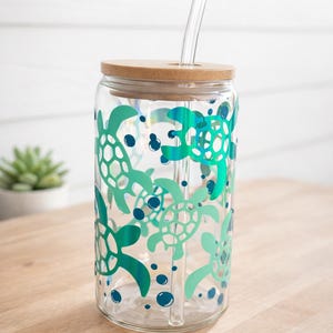 May include: A clear glass tumbler with a bamboo lid and a clear straw. The tumbler is decorated with a pattern of green and teal sea turtles and blue bubbles. The tumbler is sitting on a wooden surface.