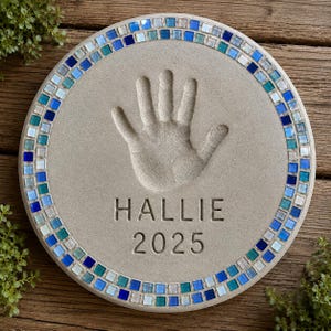 May include: A circular concrete stepping stone featuring a central handprint impression, the name HALLIE, and the year 2025 engraved in the center. The border is decorated with a mosaic of small square glass tiles in shades of blue and teal.