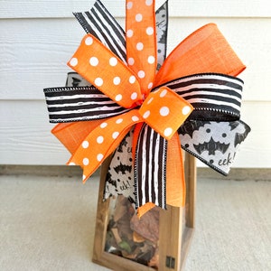 May include: A wooden lantern with a glass front is decorated with a large orange and black bow. The bow is made of various ribbons with polka dots, stripes, and a bat pattern. The lantern is filled with decorative items and has the word "eek!" written on it.