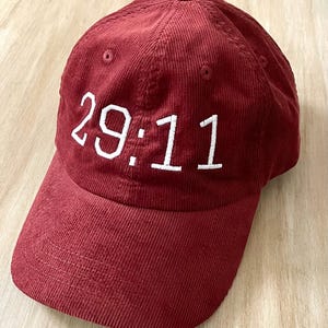 May include: A maroon corduroy baseball cap featuring the embroidered white text 29:11 on the front panel. The hat has a structured crown, a curved brim with horizontal stitching, and a button at the top centre of the six-panel construction.
