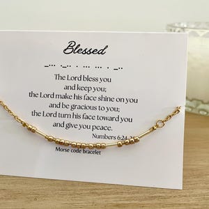 Puede incluir: Un brazalete de cuentas doradas con una tarjeta que dice "Blessed" y un versículo bíblico, "Que el Señor te bendiga y te guarde; que el Señor haga brillar su rostro sobre ti y te muestre su favor; que el Señor vuelva su rostro hacia ti y te conceda paz. Números 6:24-26." La tarjeta también dice "Brazalete de código Morse".