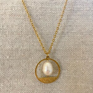 May include: A gold chain necklace with a circular pendant featuring a white pearl. The pendant has a textured, brushed gold finish.
