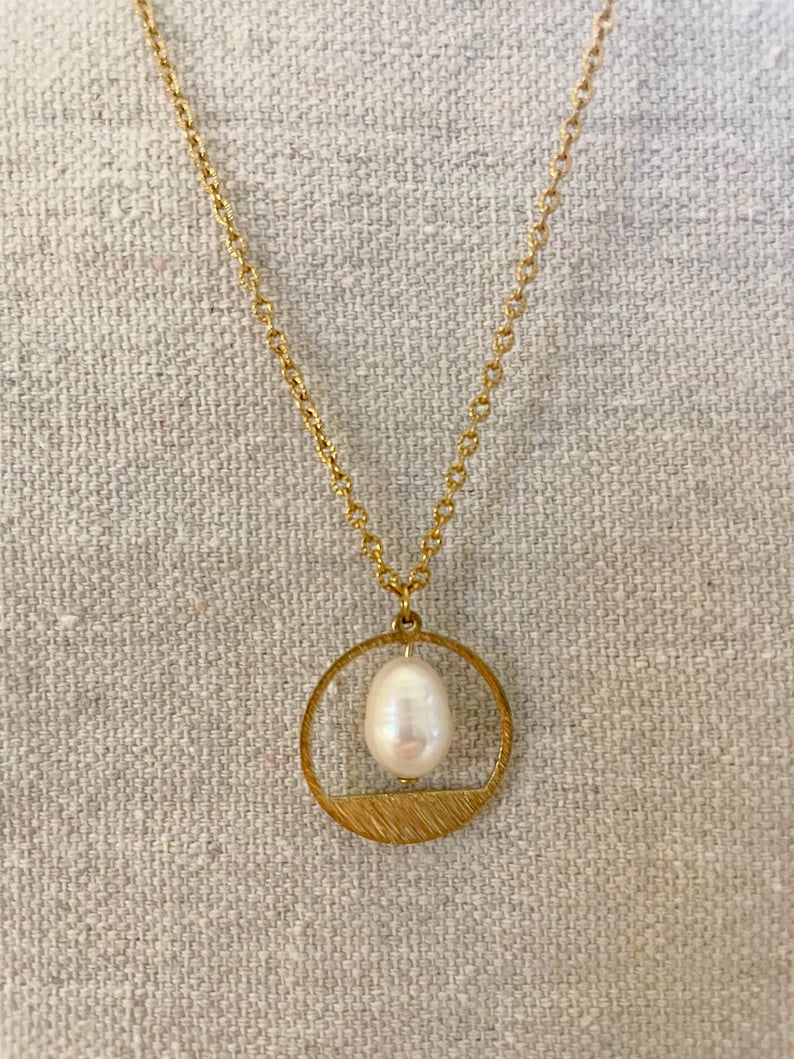 May include: A gold chain necklace with a circular pendant featuring a white pearl. The pendant has a textured, brushed gold finish.