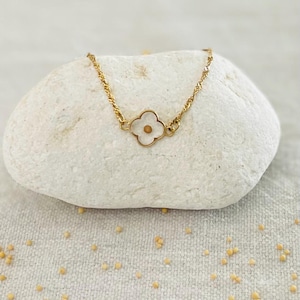 May include: A gold-toned chain bracelet featuring a central four-leaf clover charm. The charm has a white, translucent, petal-like border surrounding a small, circular, gold-colored center. The chain consists of twisted, reflective metal links.