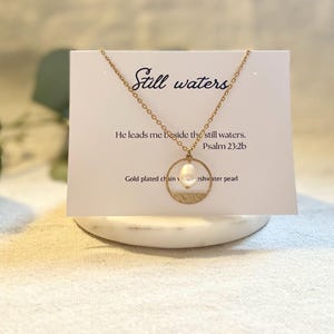 May include: Gold necklace with a freshwater pearl pendant and a circular charm with the text "Still waters" and the Bible verse "He leads me beside the still waters. Psalm 23:2b".