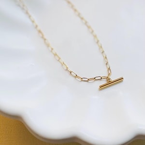 May include: A delicate gold-colored necklace with a bar pendant. The necklace features a chain with small, rectangular links. The bar pendant is horizontal and centered. The necklace is displayed on a white, scalloped surface.