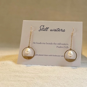 May include: Gold plated earrings with a freshwater pearl and a circle design. The earrings are hanging from a gold chain. The card reads "Still waters" and "He leads me beside the still waters. Psalm 23:2b".