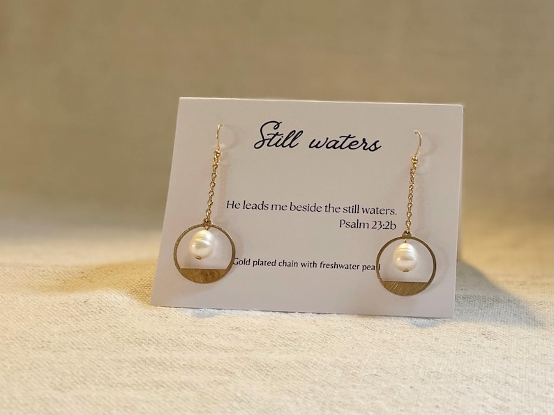 May include: Gold plated earrings with a freshwater pearl and a circle design. The earrings are hanging from a gold chain. The card reads "Still waters" and "He leads me beside the still waters. Psalm 23:2b".