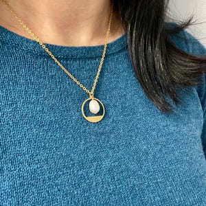 May include: A gold chain necklace with a delicate circle pendant featuring a white pearl. The necklace is worn by a person wearing a teal sweater.