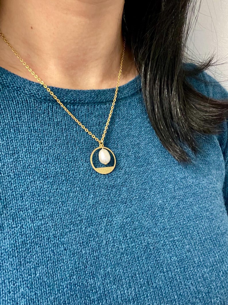 May include: A gold chain necklace with a delicate circle pendant featuring a white pearl. The necklace is worn by a person wearing a teal sweater.