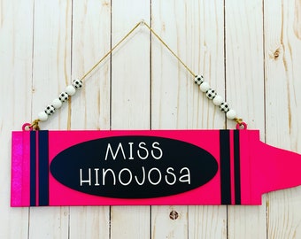 May include: Pink wooden crayon-shaped sign with black stripes and a black oval with white text that reads "Miss Hinojosa". The sign is hanging from a string with white and black checkered beads.