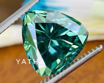 May include: A triangular green gemstone with a faceted cut, held by tweezers. The gemstone has a deep green color with a slight blue hue. The gemstone is set against a brown background.