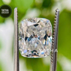 May include: A clear, cushion-cut diamond gemstone held by tweezers against a green background. The diamond has many facets and reflects light.