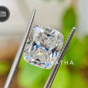 May include: A clear, faceted, cushion-cut gemstone held by a pair of tweezers. The gemstone is reflecting light and has a brilliant sparkle.