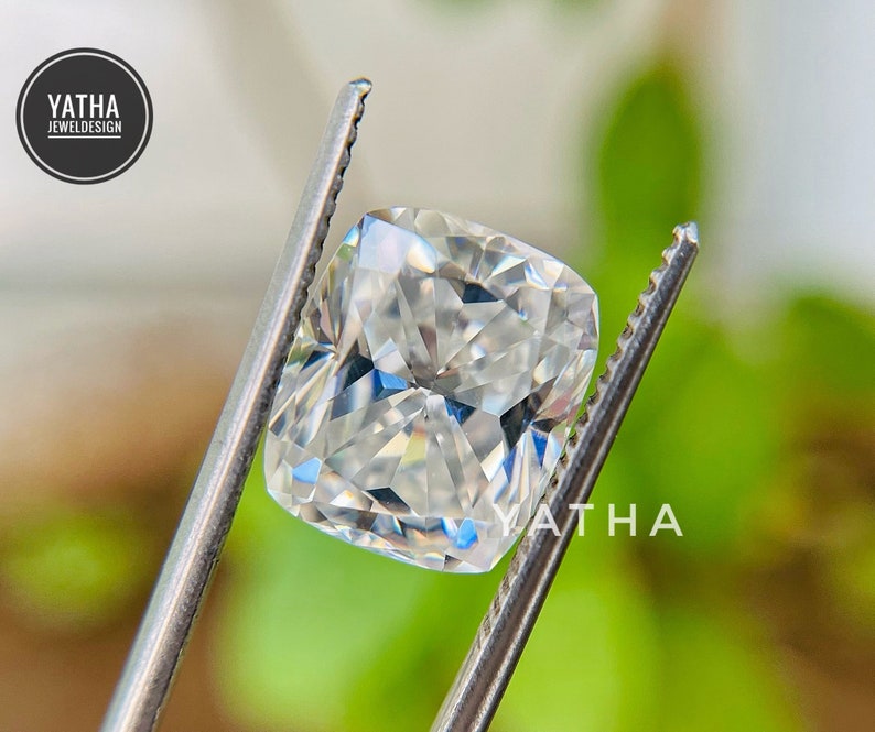 May include: A clear, faceted, cushion-cut gemstone held by a pair of tweezers. The gemstone is reflecting light and has a brilliant sparkle.