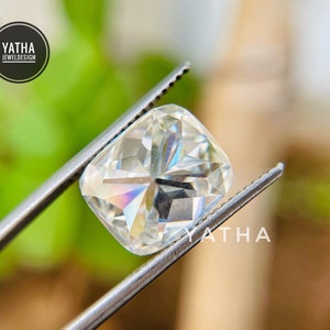 May include: A clear, square-shaped gemstone with a cushion cut, held by tweezers. The gemstone has a slight rainbow effect.