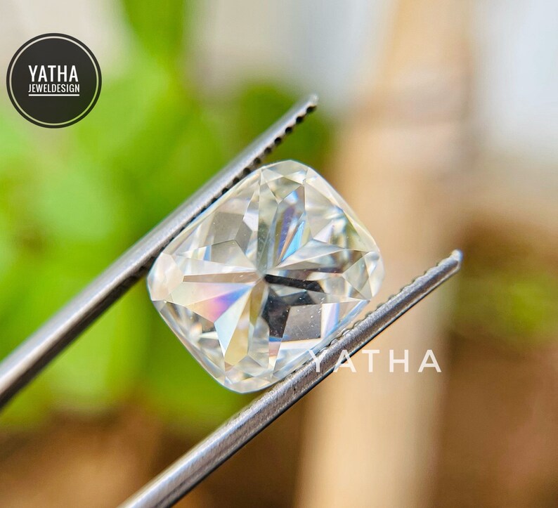 May include: A clear, square-shaped gemstone with a cushion cut, held by tweezers. The gemstone has a slight rainbow effect.