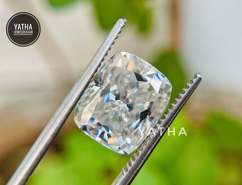 May include: A clear, cushion-cut gemstone held by tweezers. The gemstone is a brilliant white with many facets.