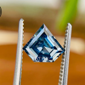 Op de afbeelding: Een blauwe vierkante edelsteen met een unieke snit, vastgehouden door een pincet. De edelsteen heeft een diepblauwe kleur en een briljante glans.