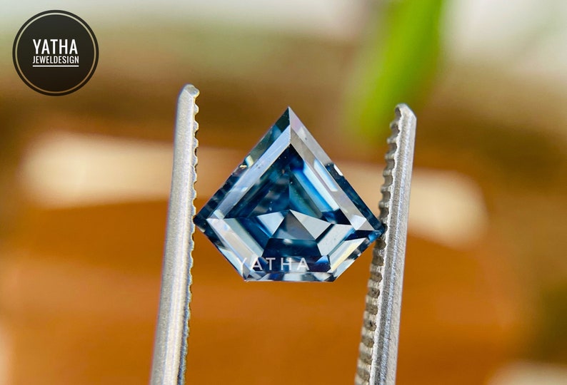 Op de afbeelding: Een blauwe vierkante edelsteen met een unieke snit, vastgehouden door een pincet. De edelsteen heeft een diepblauwe kleur en een briljante glans.