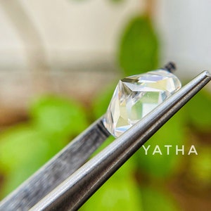May include: A clear, faceted gemstone held by tweezers against a blurred green background. The gemstone is a square shape with a slight rectangular cut. The text "YATHA" is visible in the lower right corner of the image.