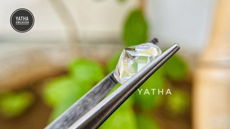 May include: A clear, faceted gemstone held by tweezers against a blurred green background. The gemstone is a square shape with a slight rectangular cut. The text "YATHA" is visible in the lower right corner of the image.