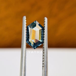 Op de afbeelding: Een blauwe edelsteen met een zeshoekige vorm, vastgehouden in een pincet. De edelsteen heeft een licht iriserende kwaliteit.