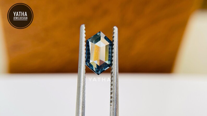 Op de afbeelding: Een blauwe edelsteen met een zeshoekige vorm, vastgehouden in een pincet. De edelsteen heeft een licht iriserende kwaliteit.