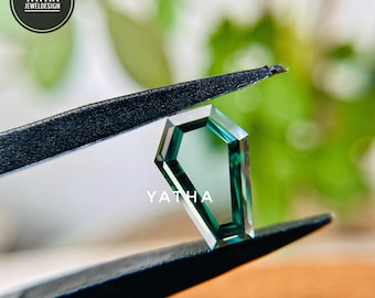 May include: A faceted, emerald-cut gemstone in shades of teal and green is held by black tweezers. The gem has a coffin-like shape. The brand name "YATHA JEWELDESIGN" is printed on the gem. The background is blurred.
