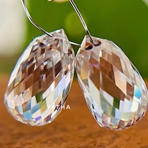 May include: A pair of clear, faceted crystal teardrop earrings. The earrings are suspended from silver-colored hooks. The crystals reflect light, displaying a spectrum of colors. The background is blurred, with a wooden surface and green foliage.
