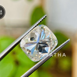 May include: A clear, cushion-cut gemstone held by tweezers. The gemstone has a brilliant, faceted surface and is set against a blurred green background.