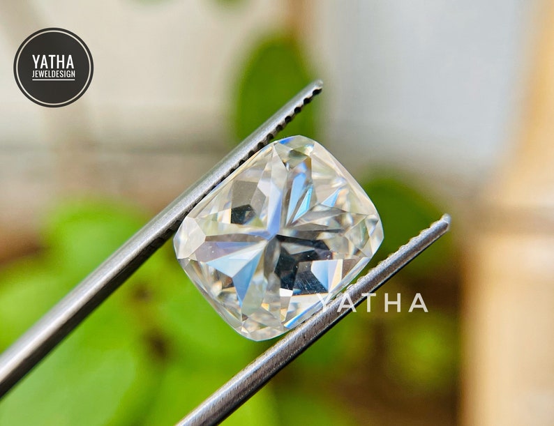 May include: A clear, cushion-cut gemstone held by tweezers. The gemstone has a brilliant, faceted surface and is set against a blurred green background.