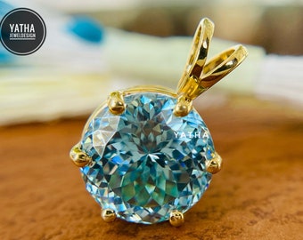 May include: A round, light blue gemstone pendant with a gold setting. The gemstone is faceted, reflecting light. The pendant has a gold bail for a chain. The logo "YATHA JEWELDESIGN" is in the upper left corner.