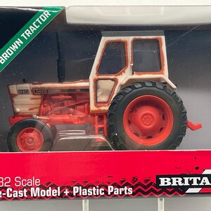 May include: A die-cast model of a brown and white David Brown tractor. The tractor has red wheels and a black tire tread. The model is labeled "1210" and is in a display box with the words "BROWN TRACTOR".