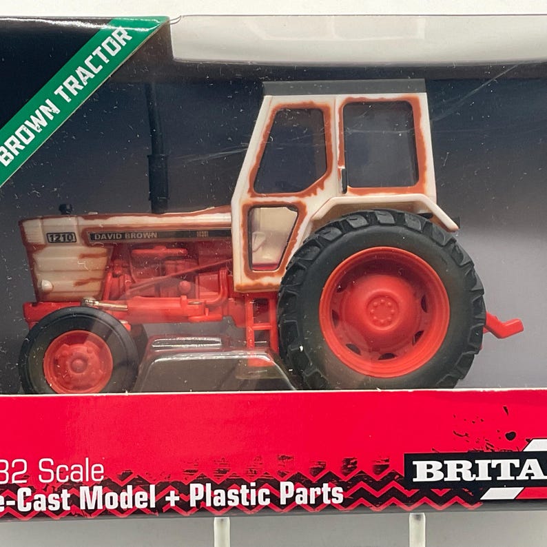 May include: A die-cast model of a brown and white David Brown tractor. The tractor has red wheels and a black tire tread. The model is labeled "1210" and is in a display box with the words "BROWN TRACTOR".