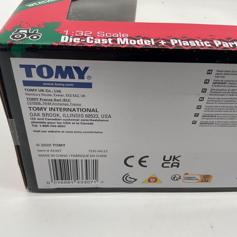 May include: A close-up of a product box with the TOMY logo and product information. The box is red, green, and white, with text that reads "1:32 Scale Die-Cast Model + Plastic Parts." The box also includes a barcode and various safety symbols.