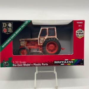 May include: A weathered David Brown tractor model in a box. The die-cast model has red and white paint, with black tires. The box is red and green with the text "WEATHERED DAVID BROWN TRACTOR" and "BRITAINS". The scale is 1:32.
