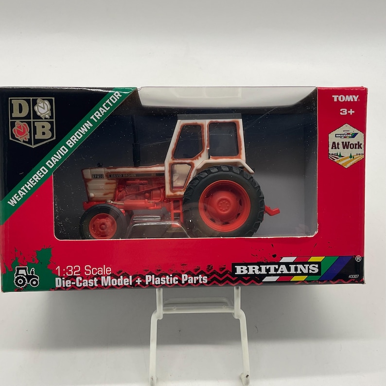 May include: A weathered David Brown tractor model in a box. The die-cast model has red and white paint, with black tires. The box is red and green with the text "WEATHERED DAVID BROWN TRACTOR" and "BRITAINS". The scale is 1:32.