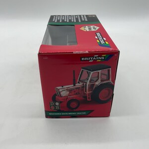 May include: A red box featuring a toy tractor. The tractor is white and red with black tires and a black roof. The box has the word "Britains" and "Weathered David Brown Tractor" printed on it.